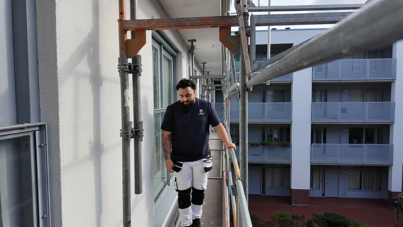 Schilder van Kwastmannen loopt over de steiger langs de galerij van een appartementencomplex, de gevel achter hem al strak wit afgewerkt.