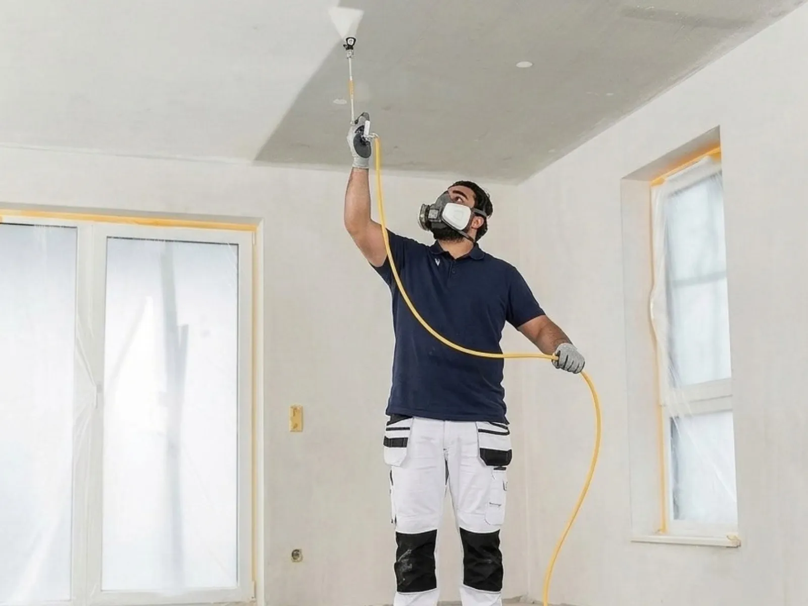 Schilder met stofmasker spuit het plafond met een airless-spuit, de ramen en kozijnen rondom afgeplakt met folie en tape.
