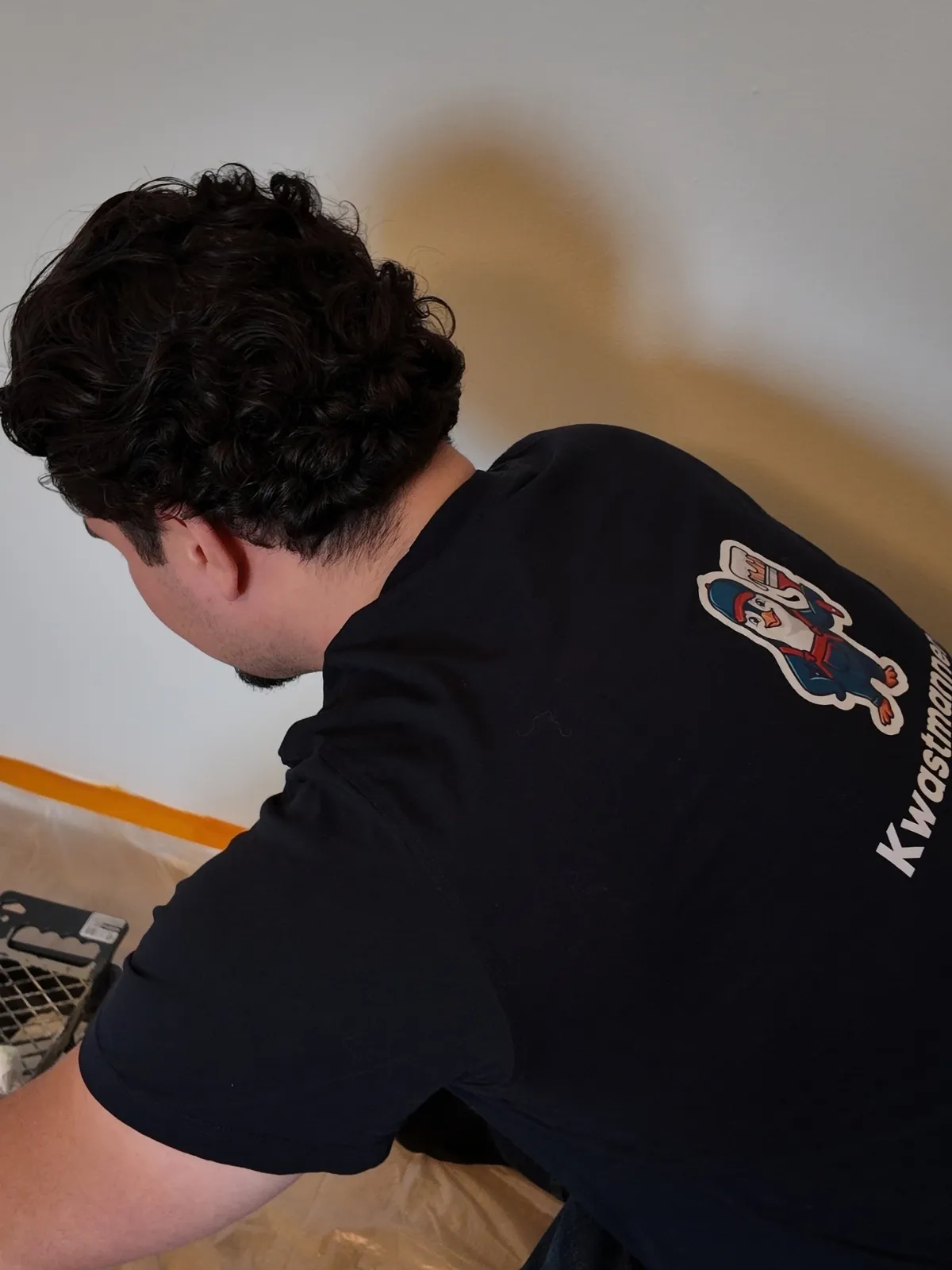 Schilder van Kwastmannen in bedrijfsshirt, gebogen over de verfbak in een kamer waar de vloer is afgedekt en de plint is afgeplakt