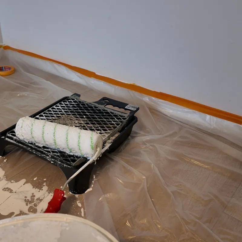Verfbak met roller op een met folie afgedekte vloer, de plint strak afgeplakt met oranje tape